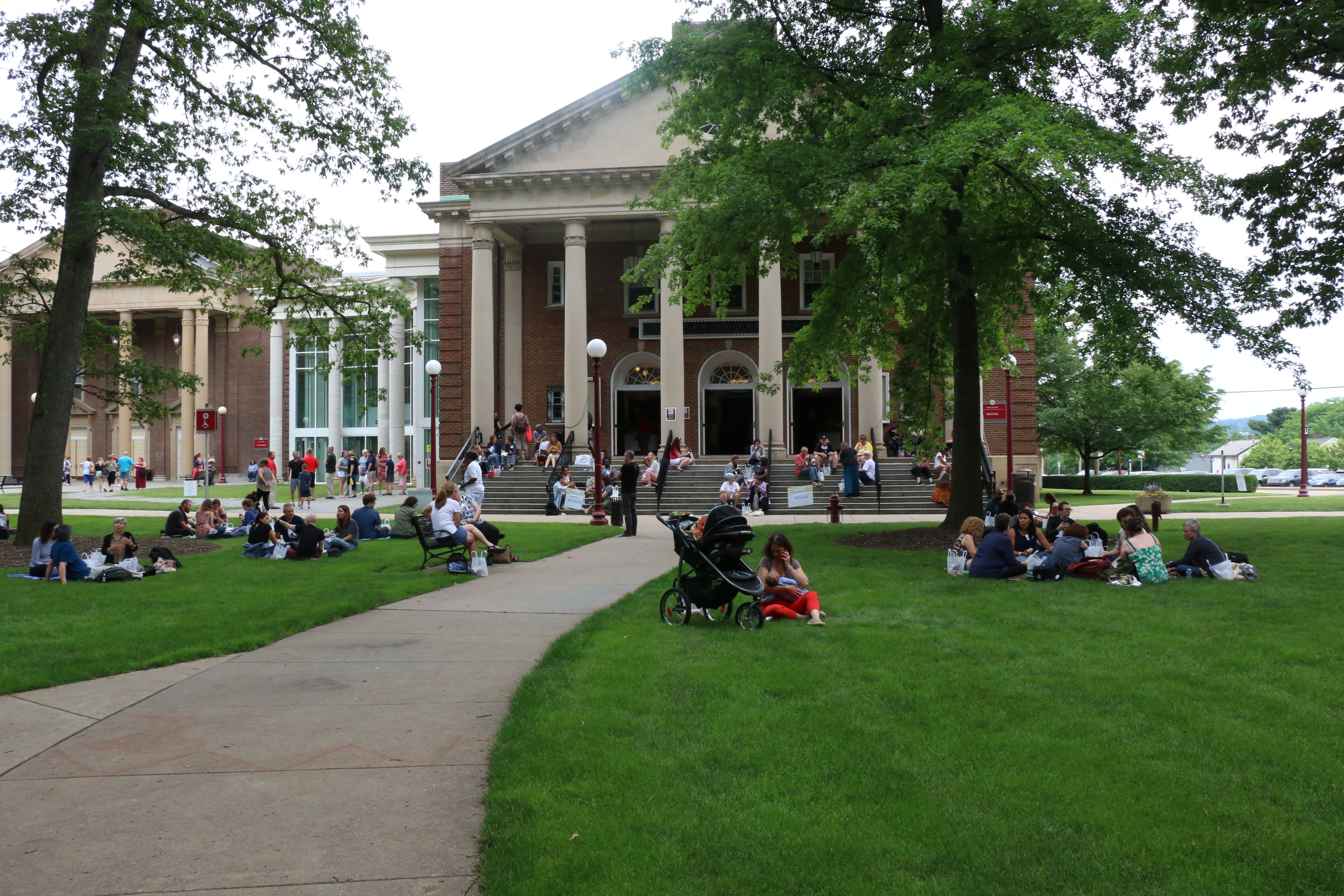 Photo of conference attendees taking a lunch break during the 2018 Conference.  Many people are sitting in the grass or on benches outside in the grassy areas surrounding the main conference building.