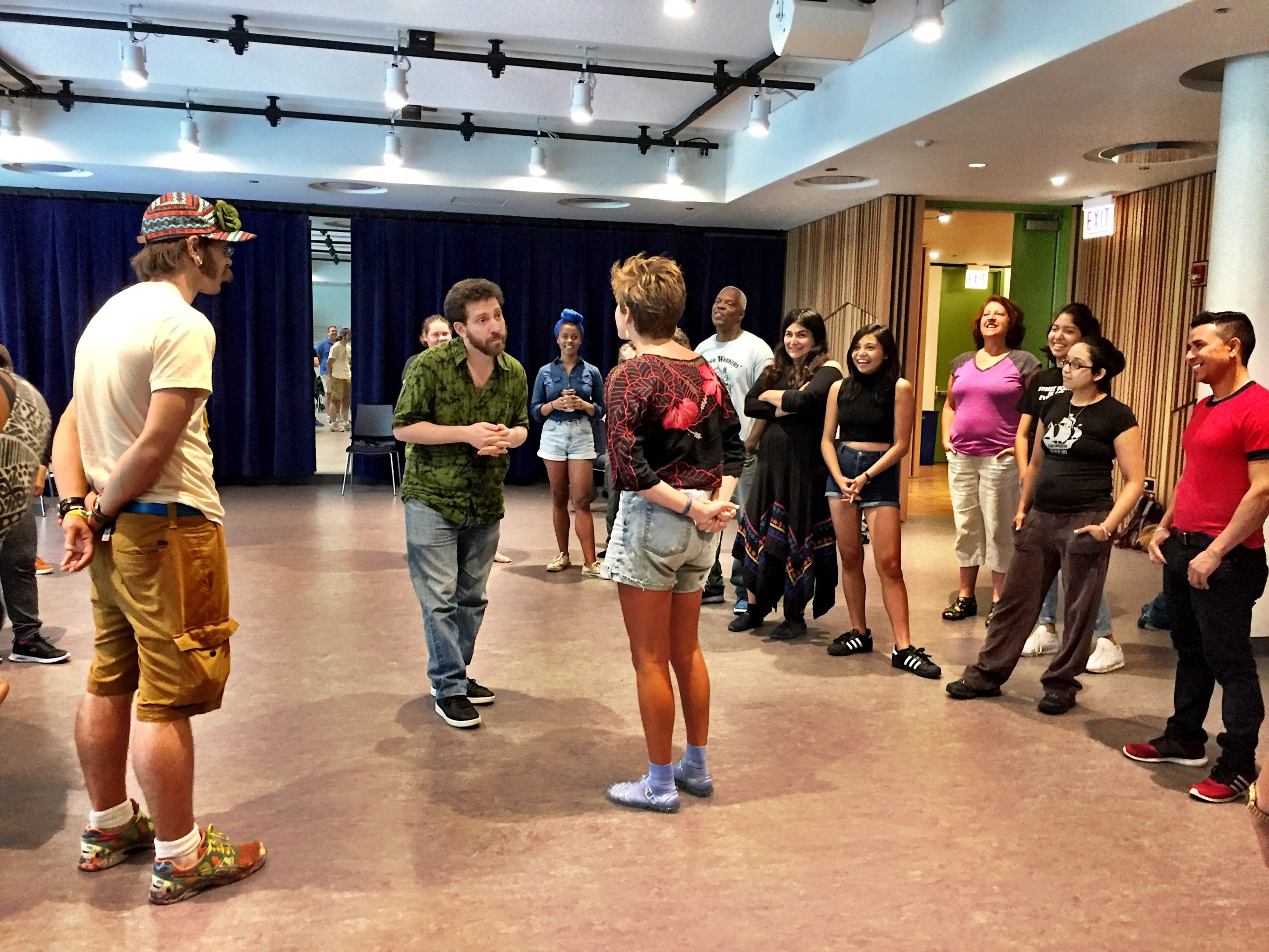 Image description: Photo of a TO workshop in which Julian Boal is jokering. Julian and a workshop participant stand in the center of a loose circle of other participants. Julian and the participant make eye contact facing each other as the other participants look on with humor.
