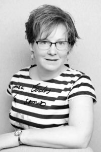 A black and white photo of Ann. She is sitting and leaning on a desk wearing a horizontally striped shirt and glasses, smiling at the camera.