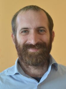 A photo of Uri from the shoulders up. He has a full beard, is wearing a light blue button up shirt without a tie, and is smiling at the camera.